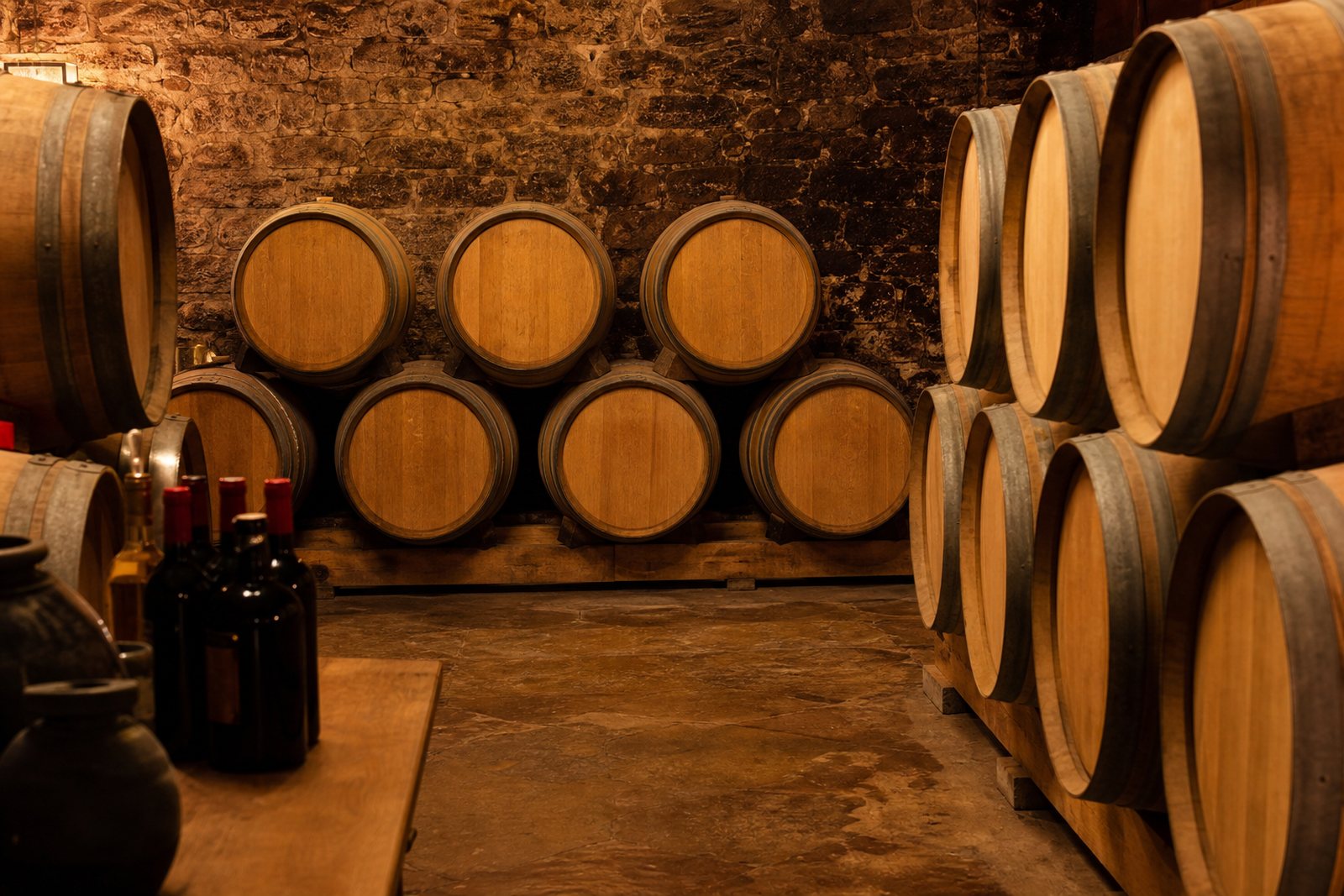 Oak wine barrels stacked in a stone cellar with a few bottles resting on a wooden table